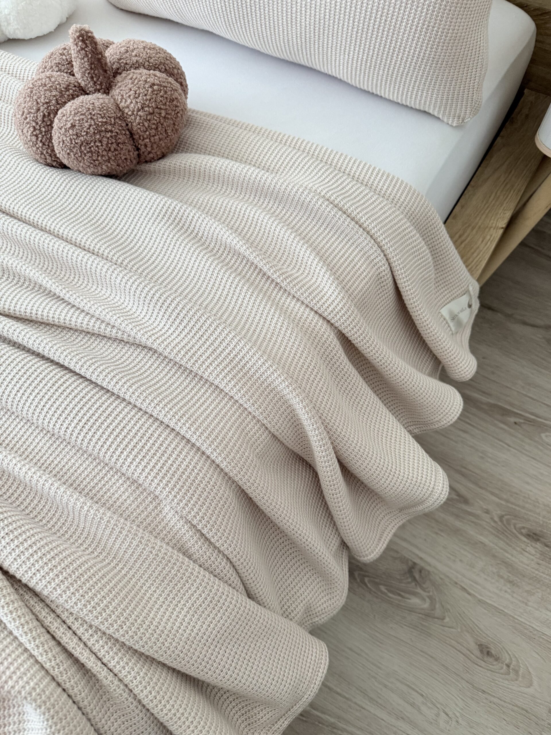 Jasny beżowy, miękki dzianinowy kocyk przewieoszony po boku łóżka idealny do salony sypialni, prezentujący dekoracyjny charakter i delikatny połysk materiału